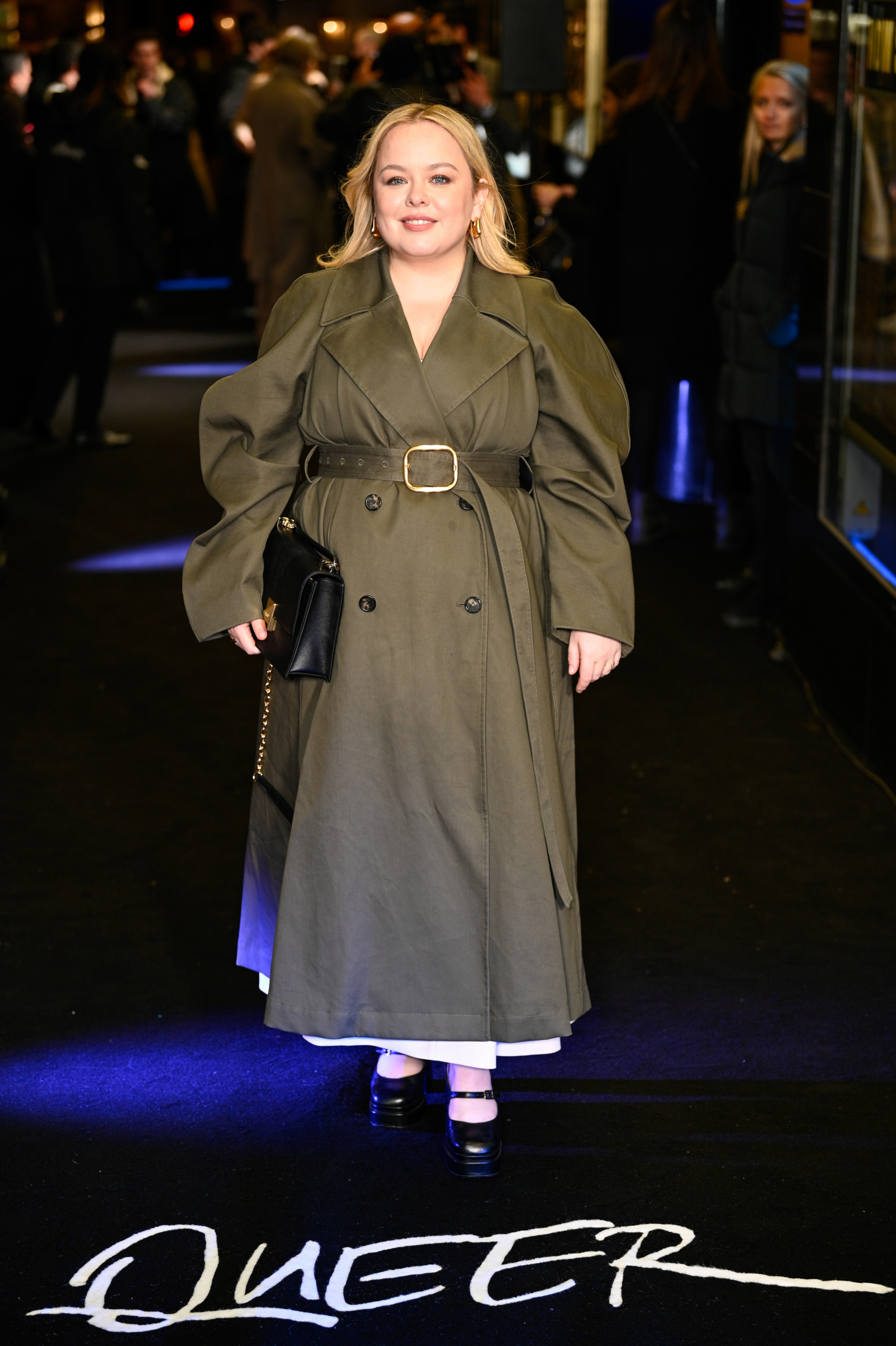 Nicola w trenczu khaki, stoi na czarnym dywanie z napisem "QUEER". Eleganckie buty dopełniają jej look.
