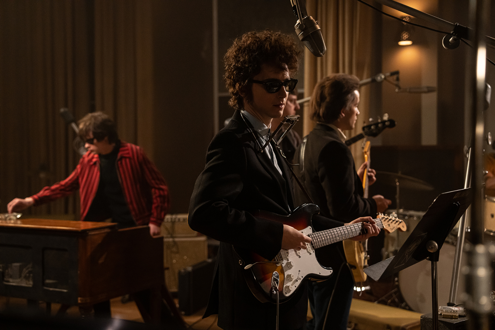 Timothée Chalamet jako Bob Dylan w filmie "Kompletnie nieznany" gra na gitarze w zespole muzycznym, podczas nagrania w studiu.