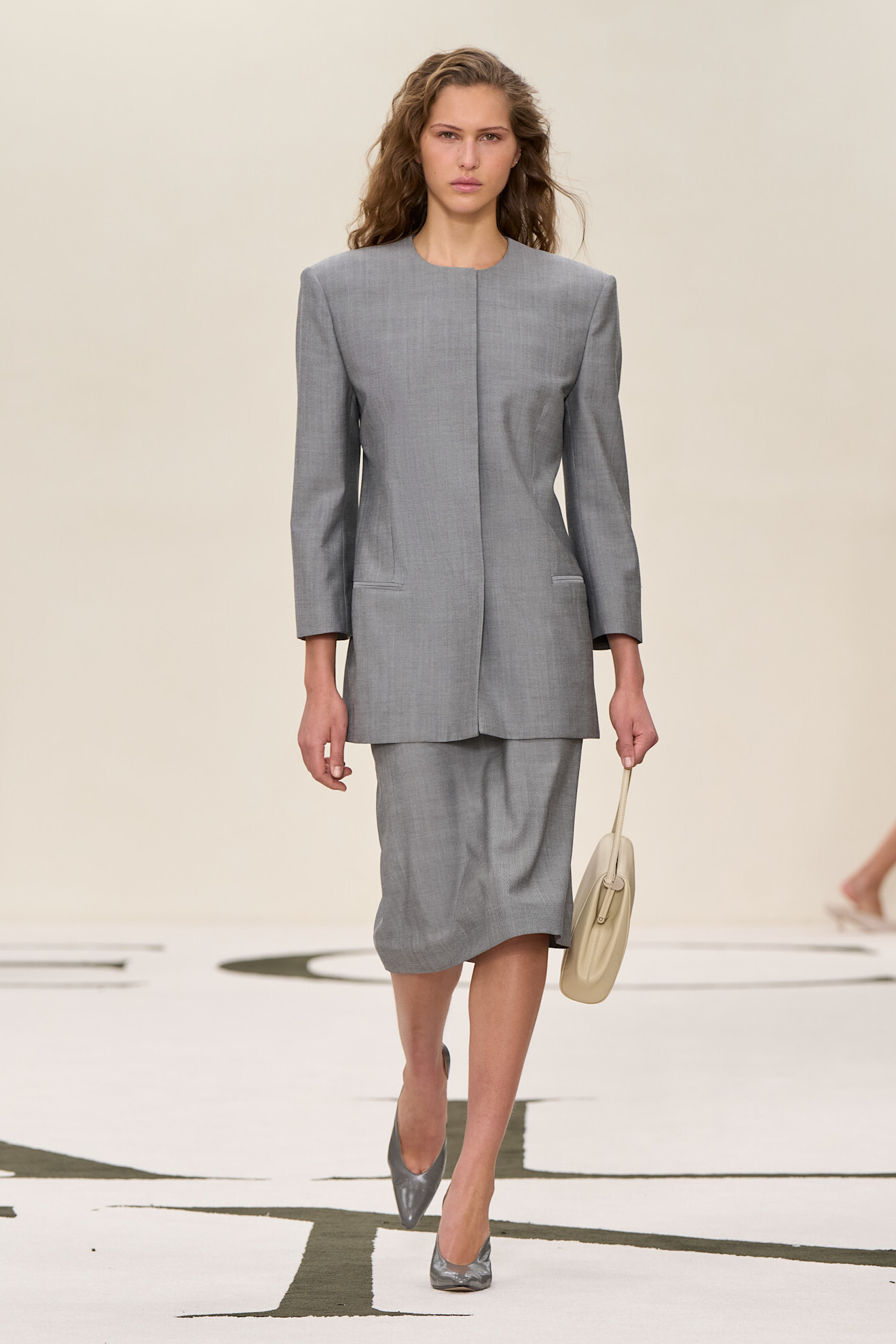 Modelka w szarym kostiumie od Calvin Klein, trzymająca torebkę w neutralnym kolorze, prezentuje elegancki styl.