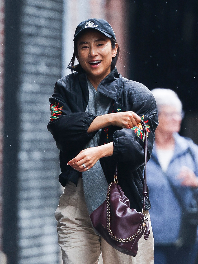 Greta Lee uśmiechnięta, w czarnej kurtce z haftem i czapce, trzyma fioletową torebkę, spacerując w mieście.
