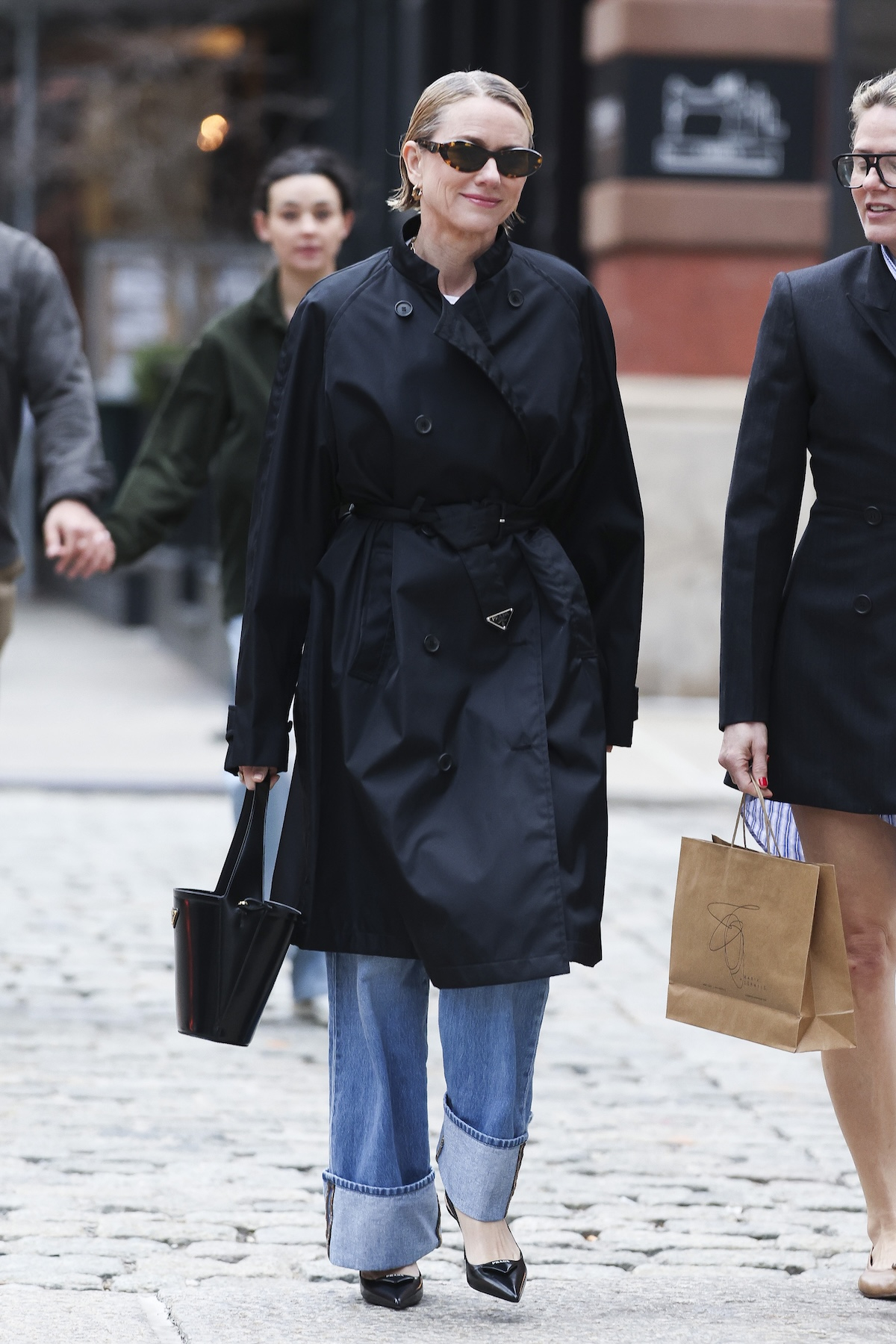 Naomi Watts w czarnym płaszczu spaceruje w jeansach z podwiniętymi nogawkami, uśmiechając się.