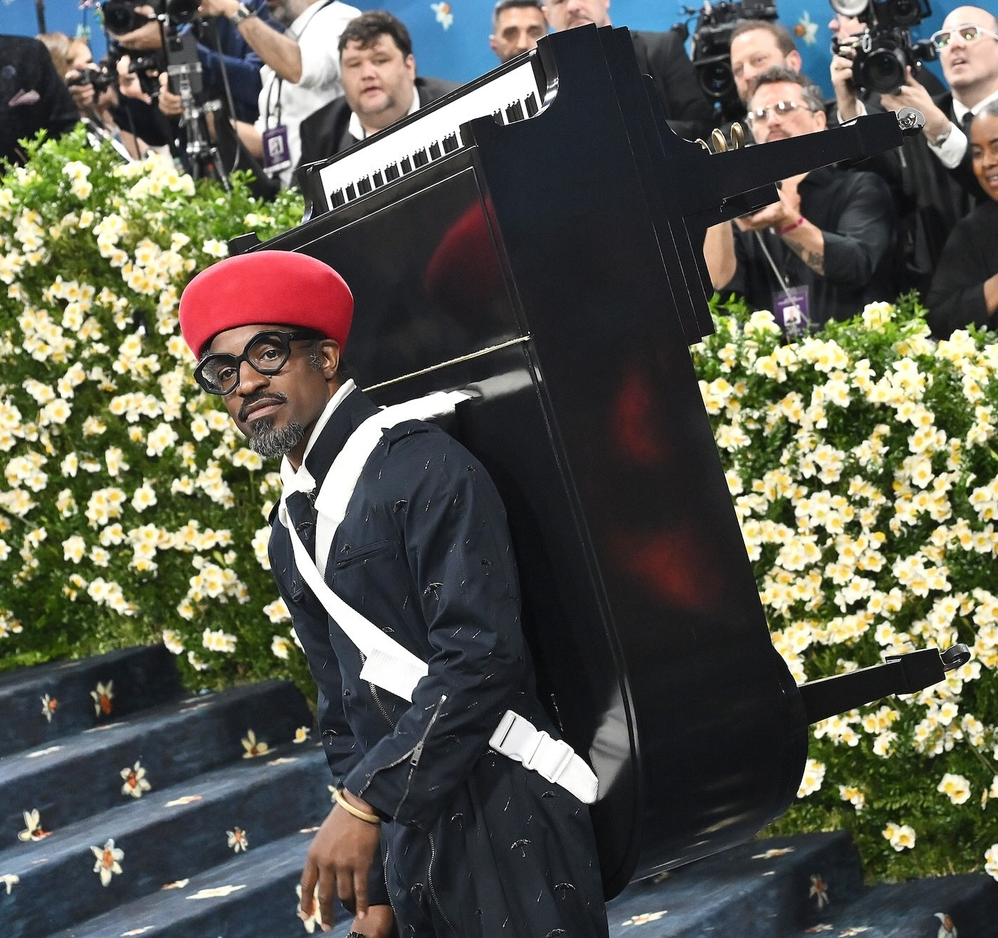 Raper Andre 3000 podczas MET Gala 2025 ma na sobie zapinany na zamki kombinezon z przetarciami, czerwony beret, złotą biżuterię, na plecach ma przyczepione za pomocą białych pasów czarne pianino z czarno-białymi klawiszami.