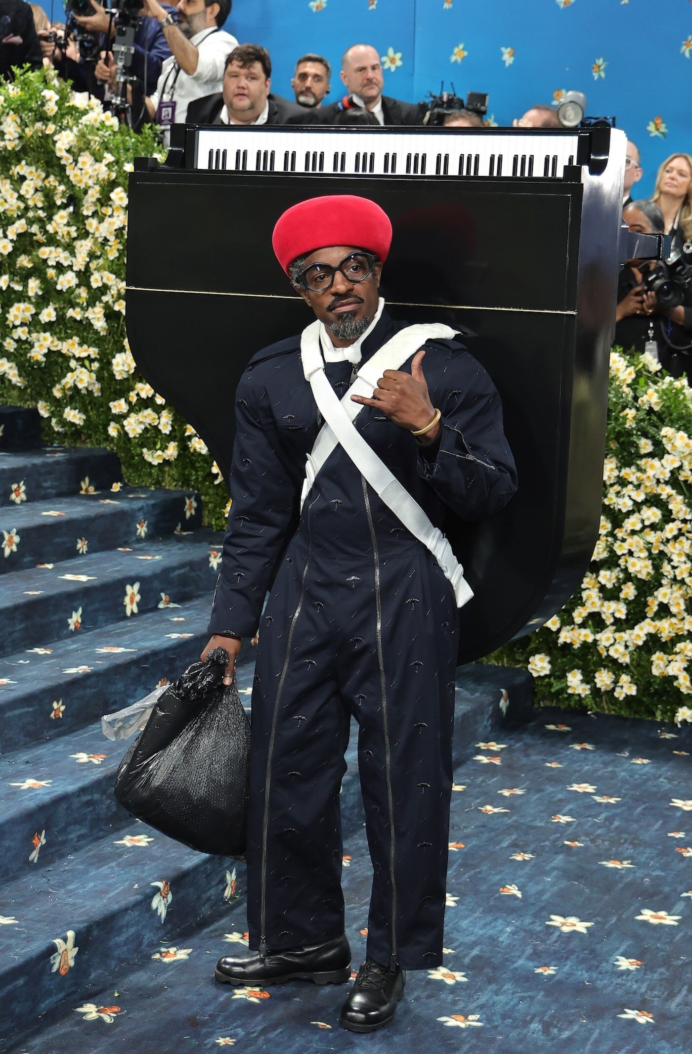 Raper Andre 3000 podczas MET Gala 2025 ma na sobie zapinany na zamki kombinezon z przetarciami, czerwony beret, czarne skórzane buty, złotą biżuterię, na plecach ma przyczepione za pomocą białych pasów czarne pianino z czarno-białymi klawiszami, w dłoni trzyma foliową torbę.