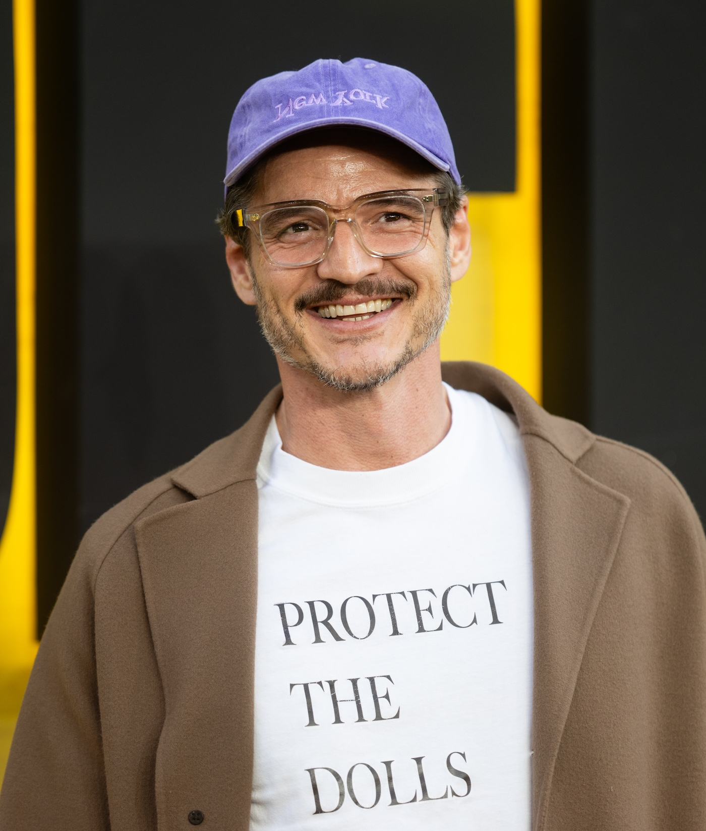 Pedro Pascal pozuje na czerwonym dywanie podczas specjalnego pokaz filmu „Thunderbolts*” w Londynie, ma na sobie biały T-shirt z nadrukiem „Protect the Dolls” projektu Connera Ivesa, długi, jasnobrązowy płaszcz i fioletową czapkę z daszkiem.