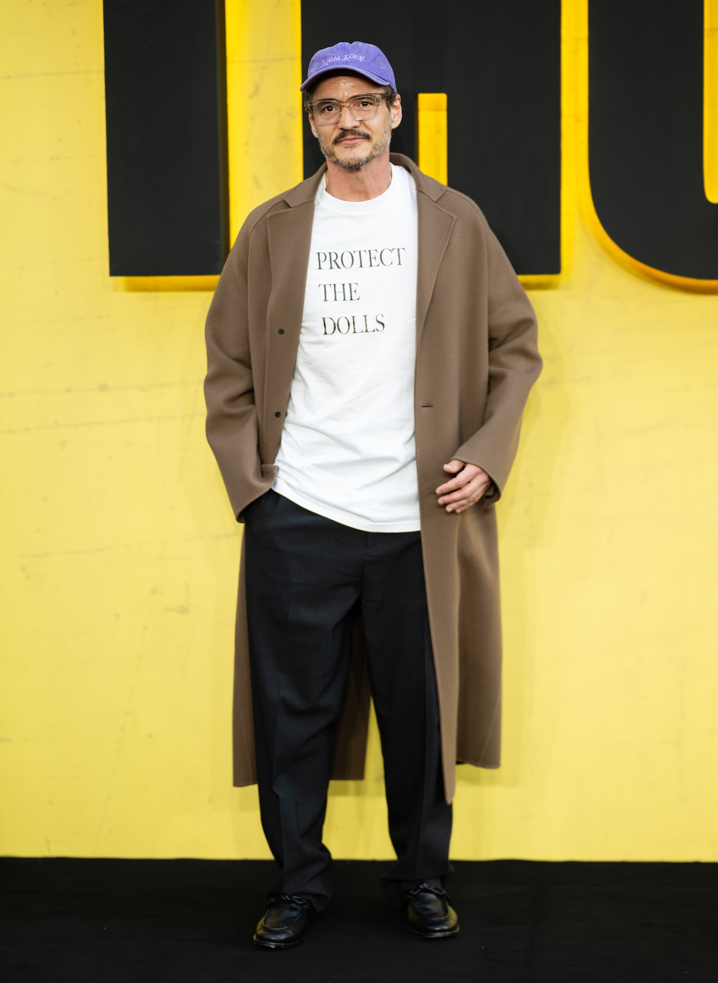 Pedro Pascal pozuje na czerwonym dywanie podczas specjalnego pokaz filmu „Thunderbolts*” w Londynie, ma na sobie biały T-shirt z nadrukiem „Protect the Dolls” projektu Connera Ivesa, długi, jasnobrązowy płaszcz, luźne, czarne spodnie zaprasowane w kant, czarne skórzane mokasyny i fioletową czapkę z daszkiem.
