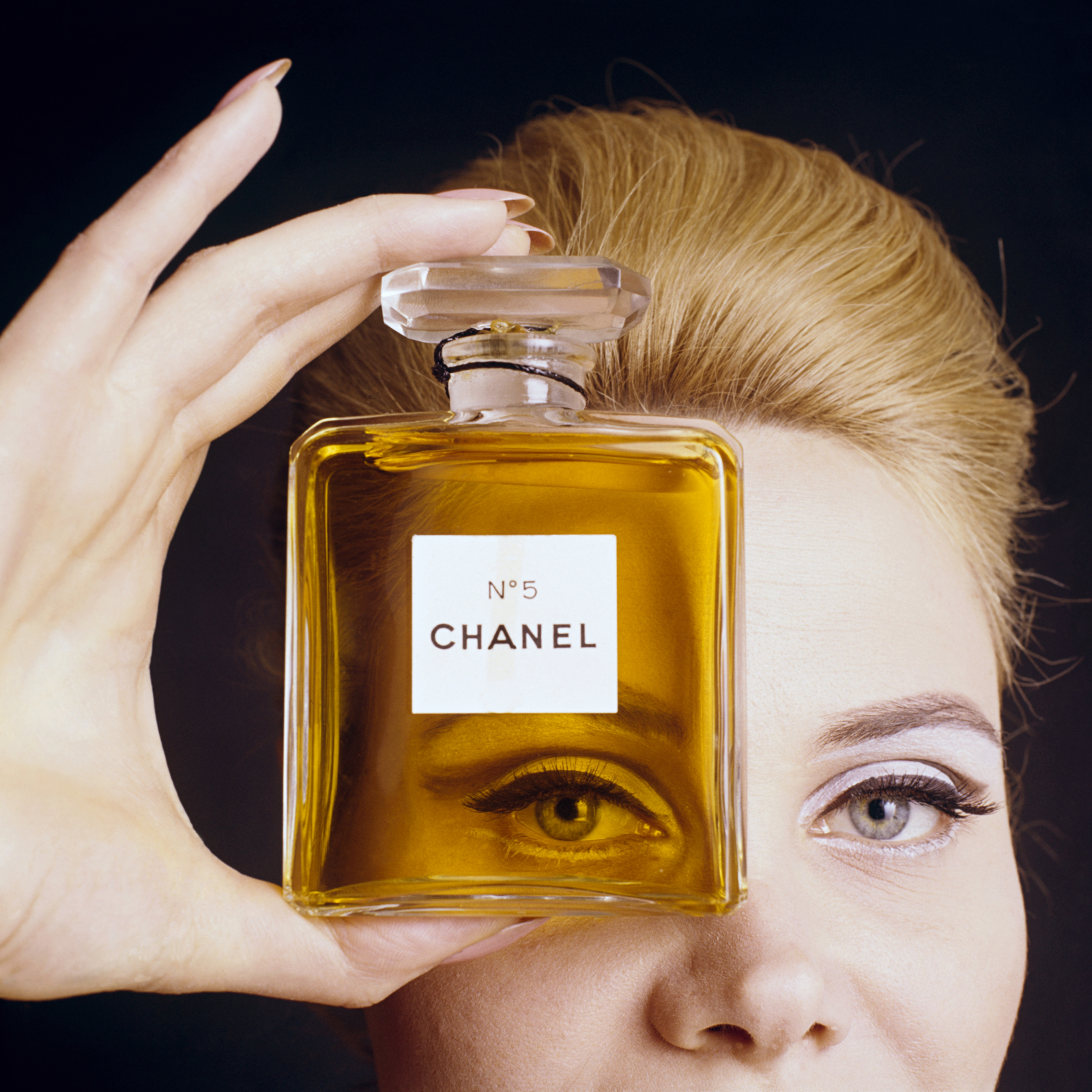 Modelka trzyma perfumy Chanel No. 5, eksponując je przed twarzą. Złota butelka z kryształowym korkiem w centralnym punkcie obrazu.