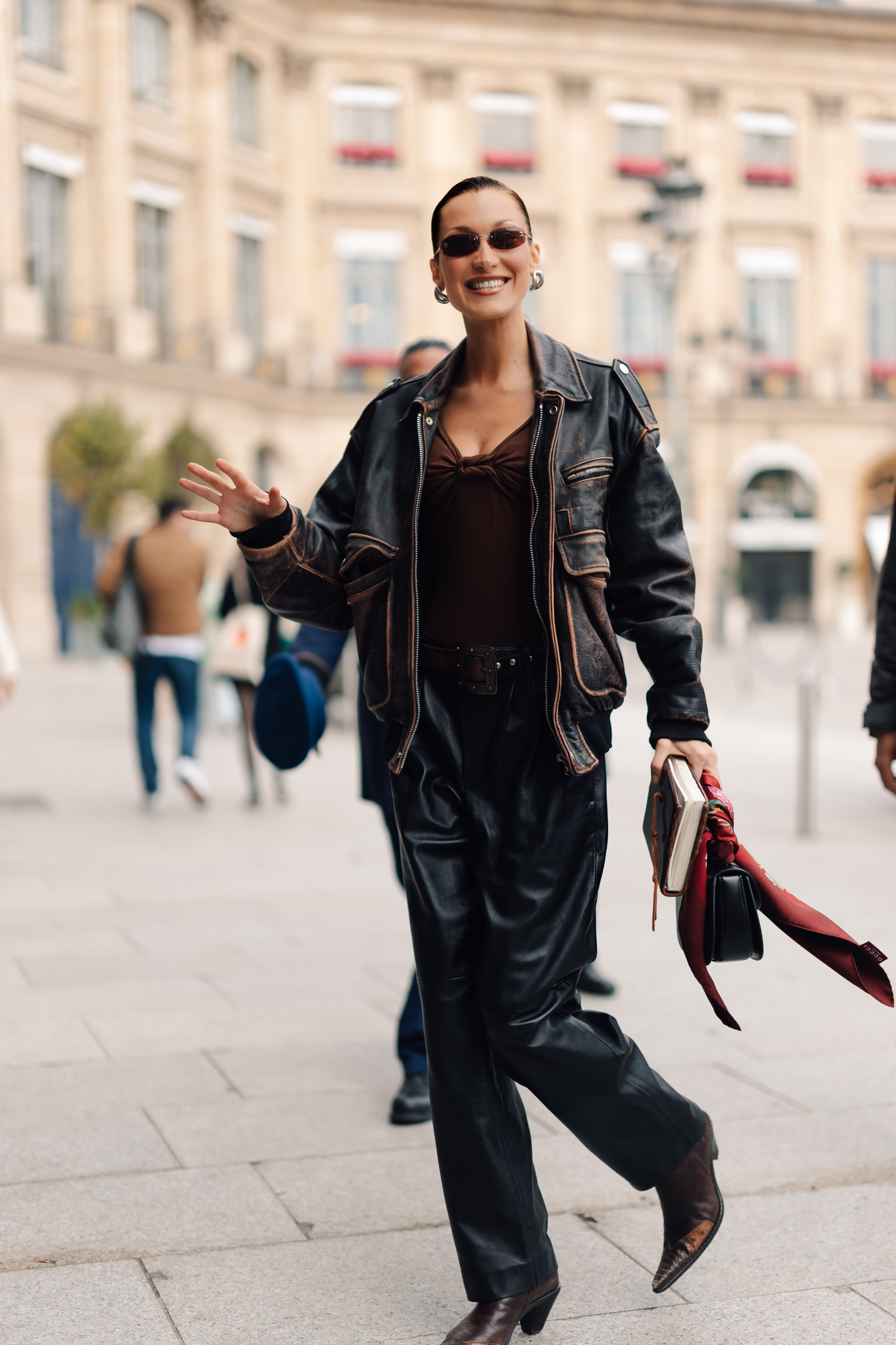 Bella Hadid w skórzanej kurtce, uśmiechając się, spaceruje po mieście w eleganckim stroju.