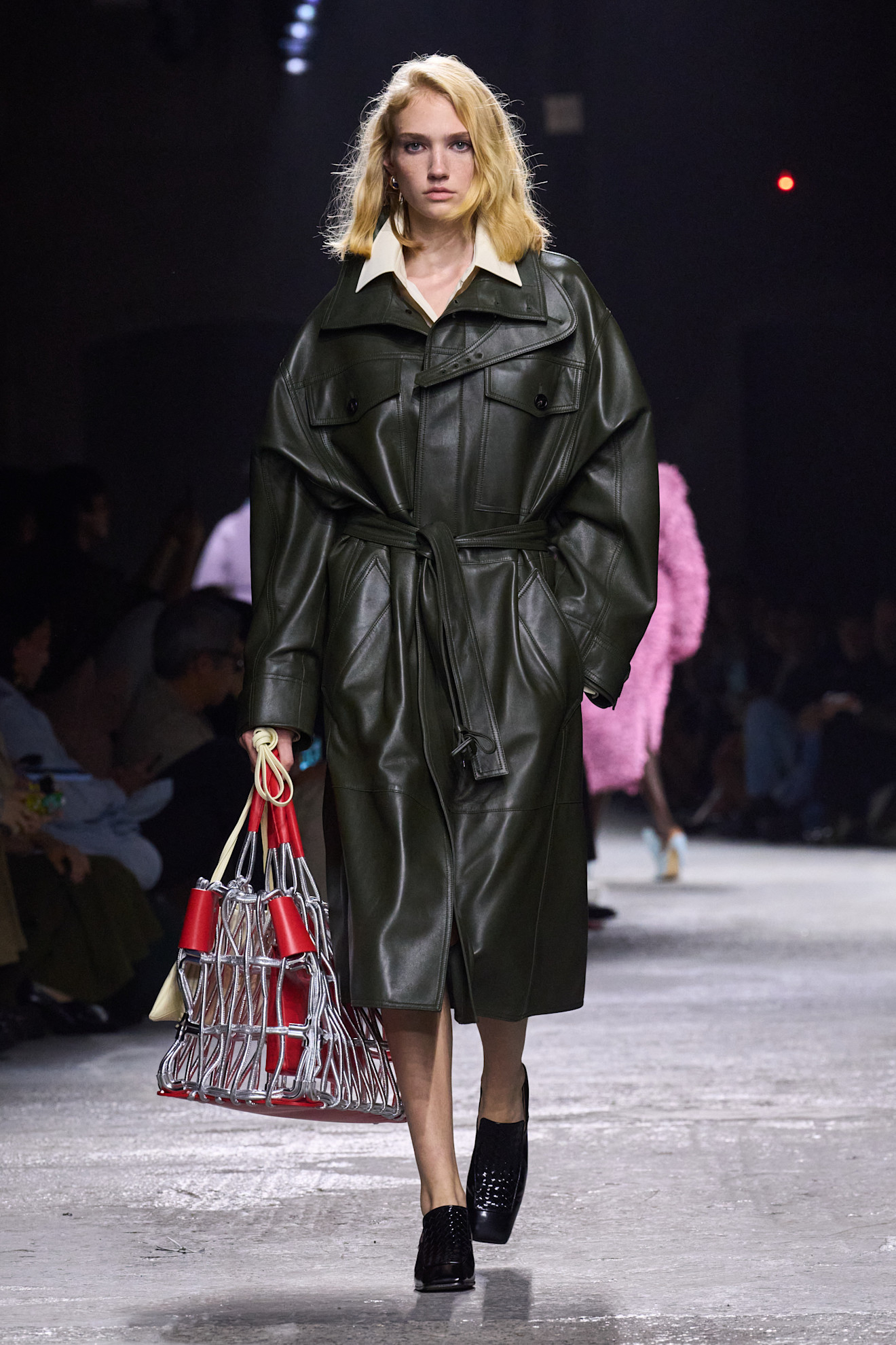 Modelka w oversize'owym, oliwkowym płaszczu idzie po wybiegu, trzymając pastelową torebkę z siatką.