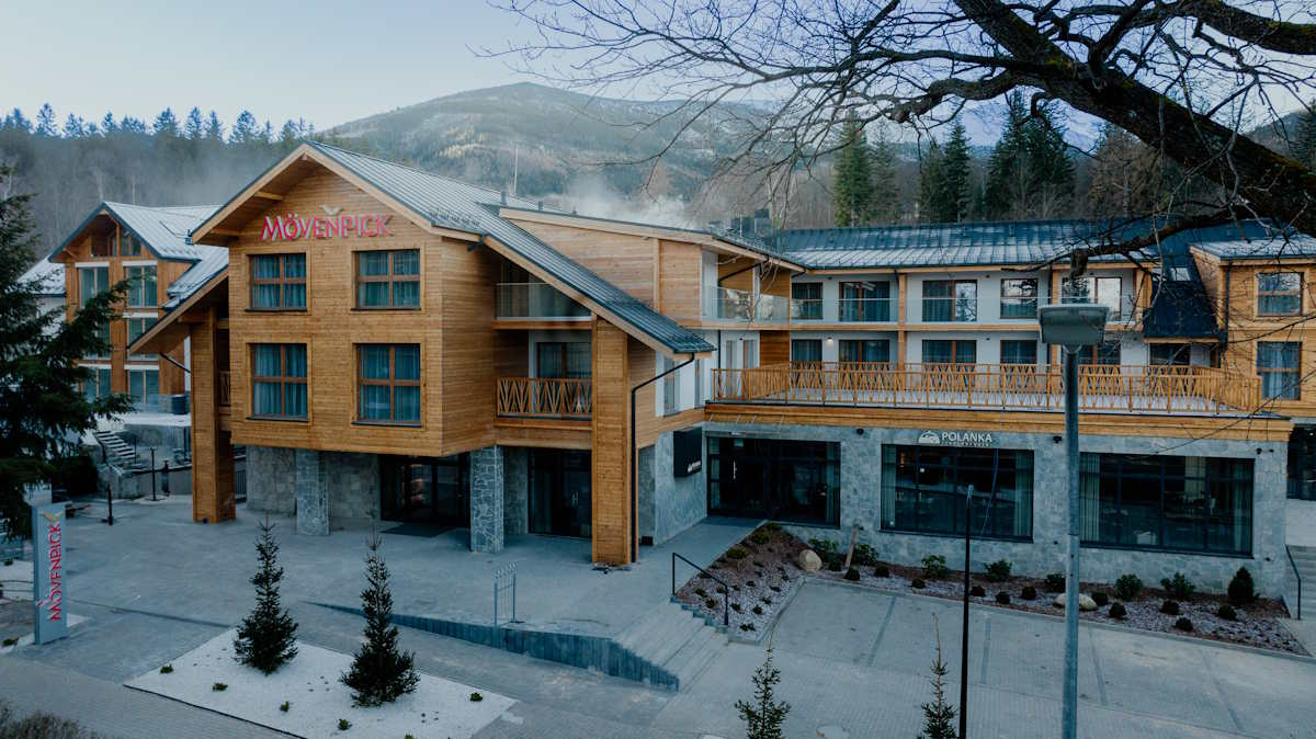 Stylowy budynek hotelu Mövenpick Resort & Spa Karpacz, otoczony drzewami i górami, z dużymi oknami i drewnianą elewacją.