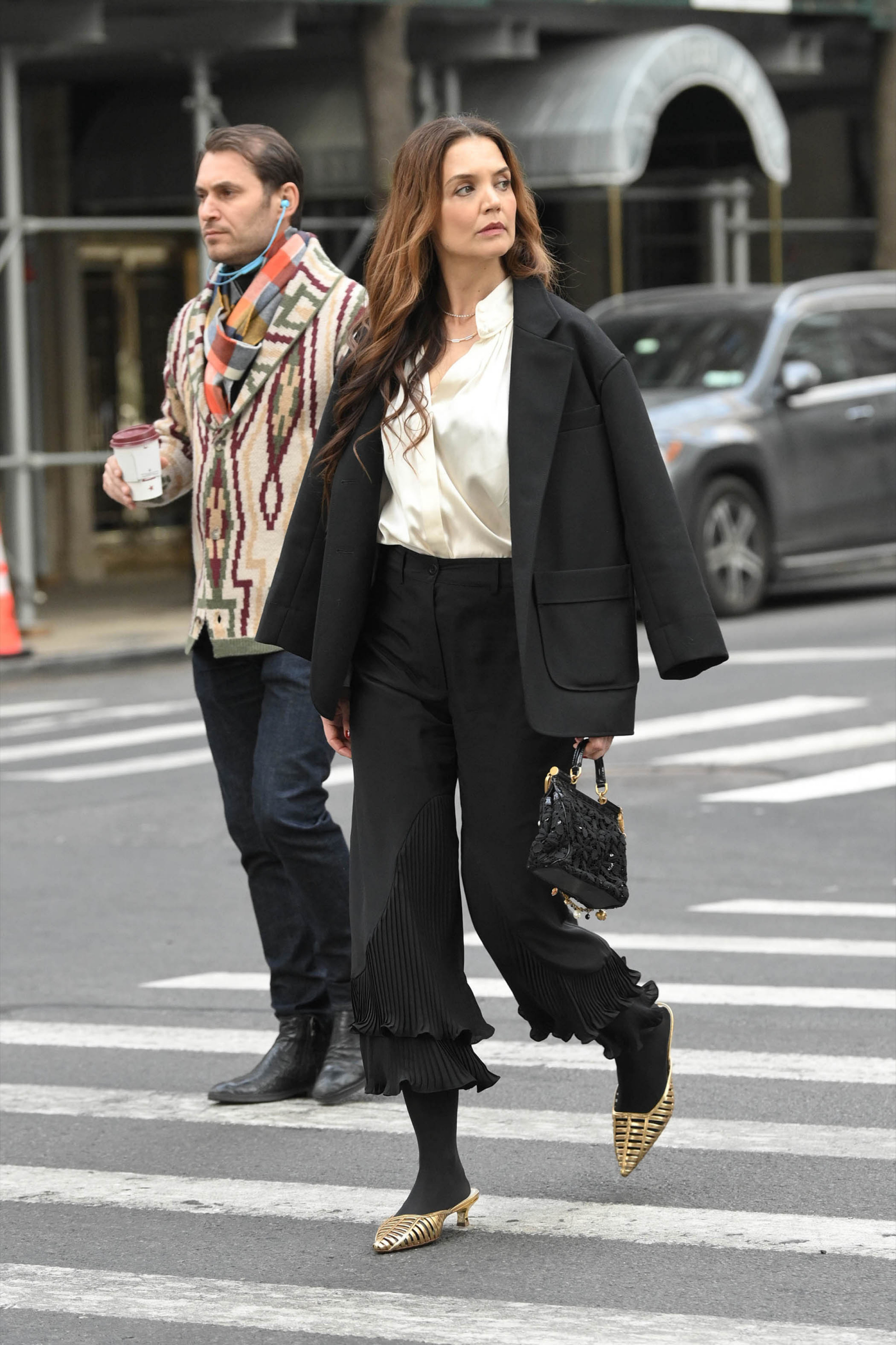 Katie Holmes w eleganckich spodniach z falbanką, czarnym żakiecie i stylowych butach, przechodzi przez ulicę.