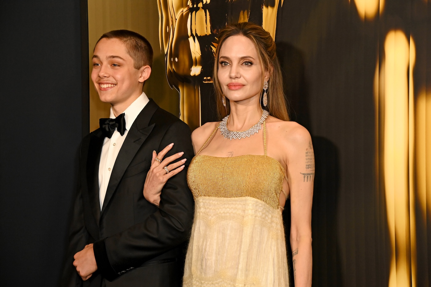 Knox Leon Jolie-Pitt i Angelina Jolie pozują razem na czerwonym dywanie, oboje ubrani w eleganckie stroje: Angelina Jolie w suknię vintage z The Knit Vintage a Knox Leon w elegancki czarny smoking, białą koszulę i muszkę.