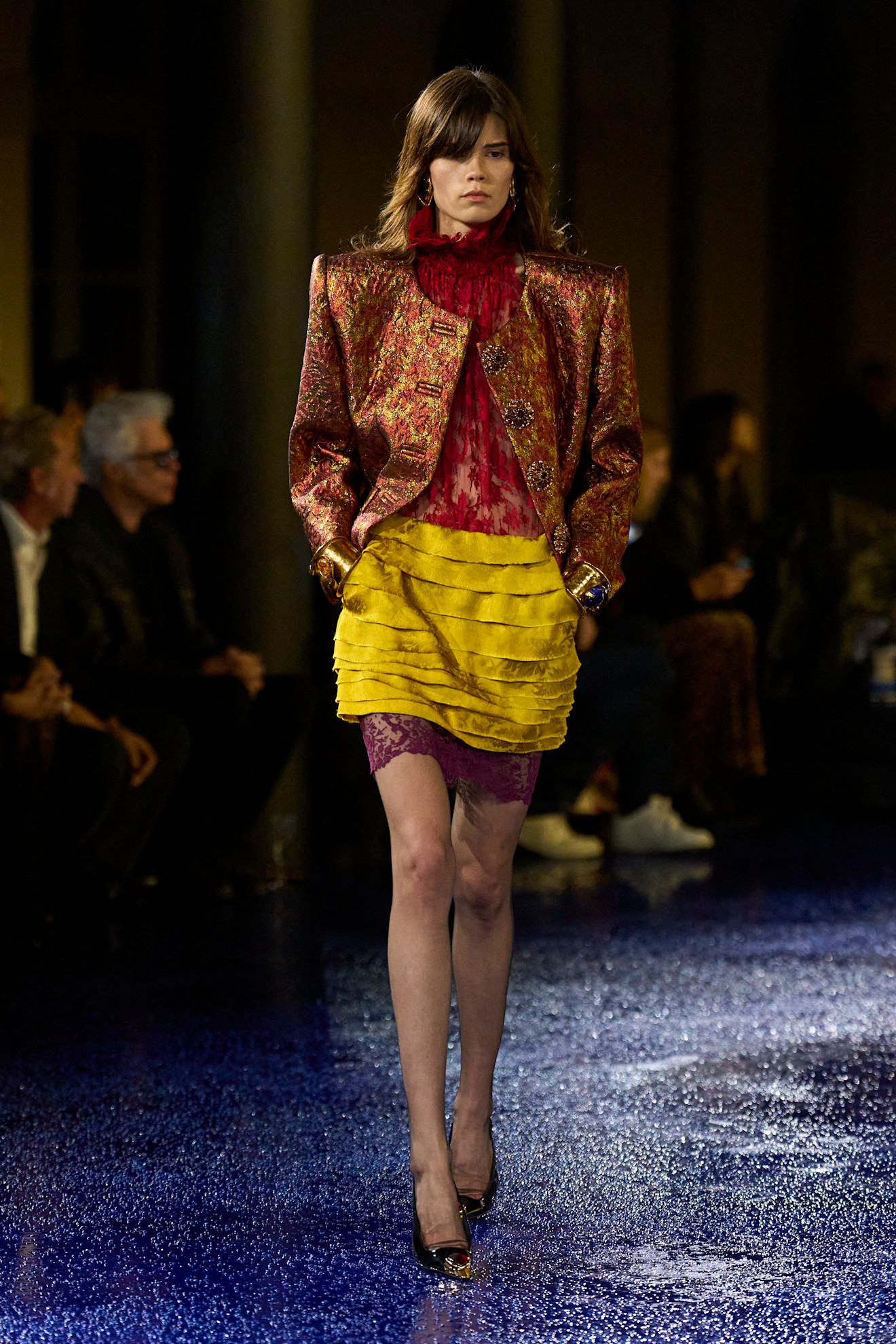 Modelka na pokazie Saint Laurent w Anja Rubik w żakiecie bez klap w stylu retro z brokatowej tkaniny, golfie z czerwonej koronki i żółtej, satynowej spódniczce z falbankami, rajstopach i czółenkach slingback z biżuteryjnym detalem.