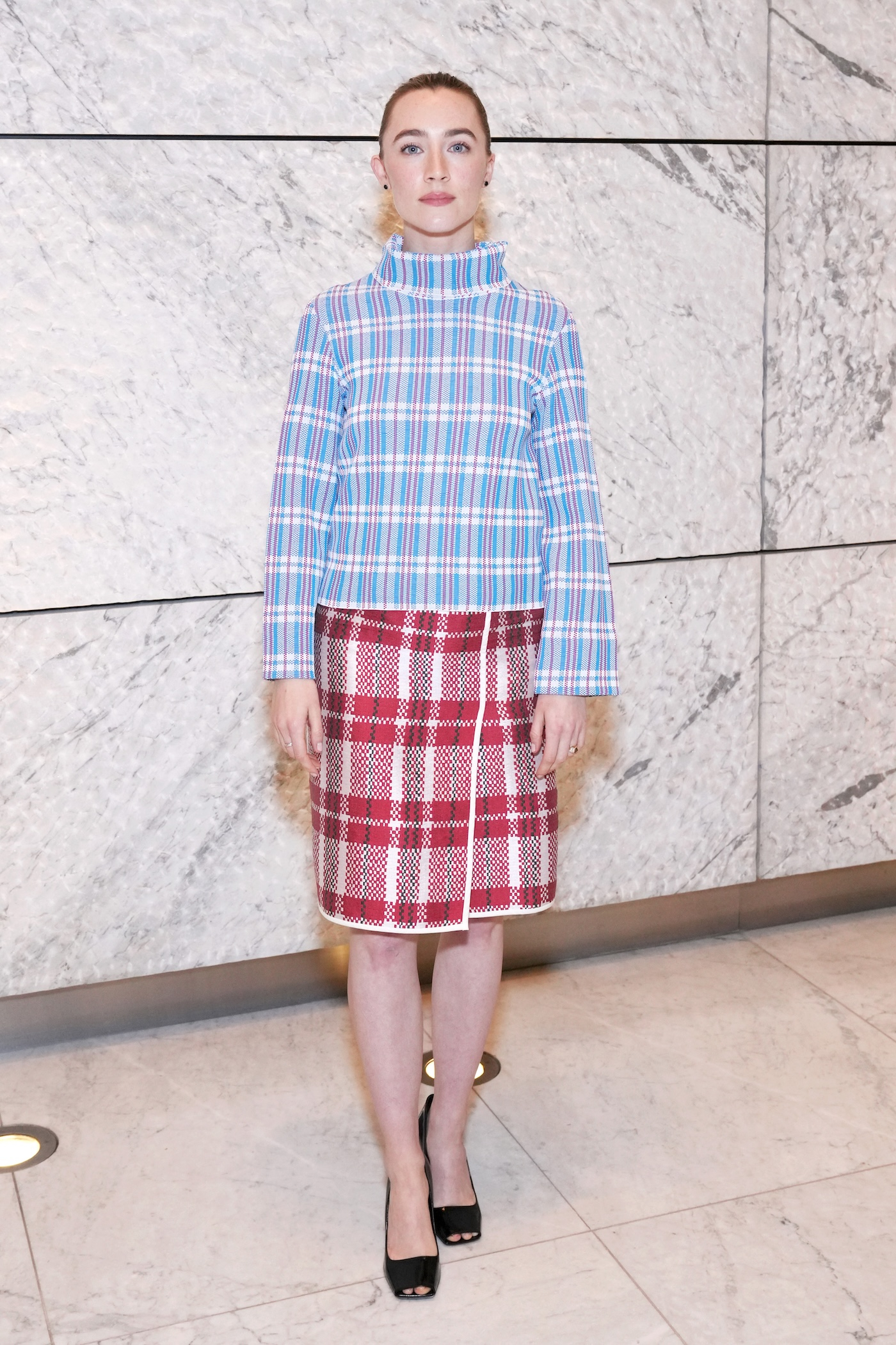 Saoirse Ronan stoi w swetrze w niebiesko-różową kratę i w czerwonej kraciastej spódnicy, na tle marmurowej ściany.