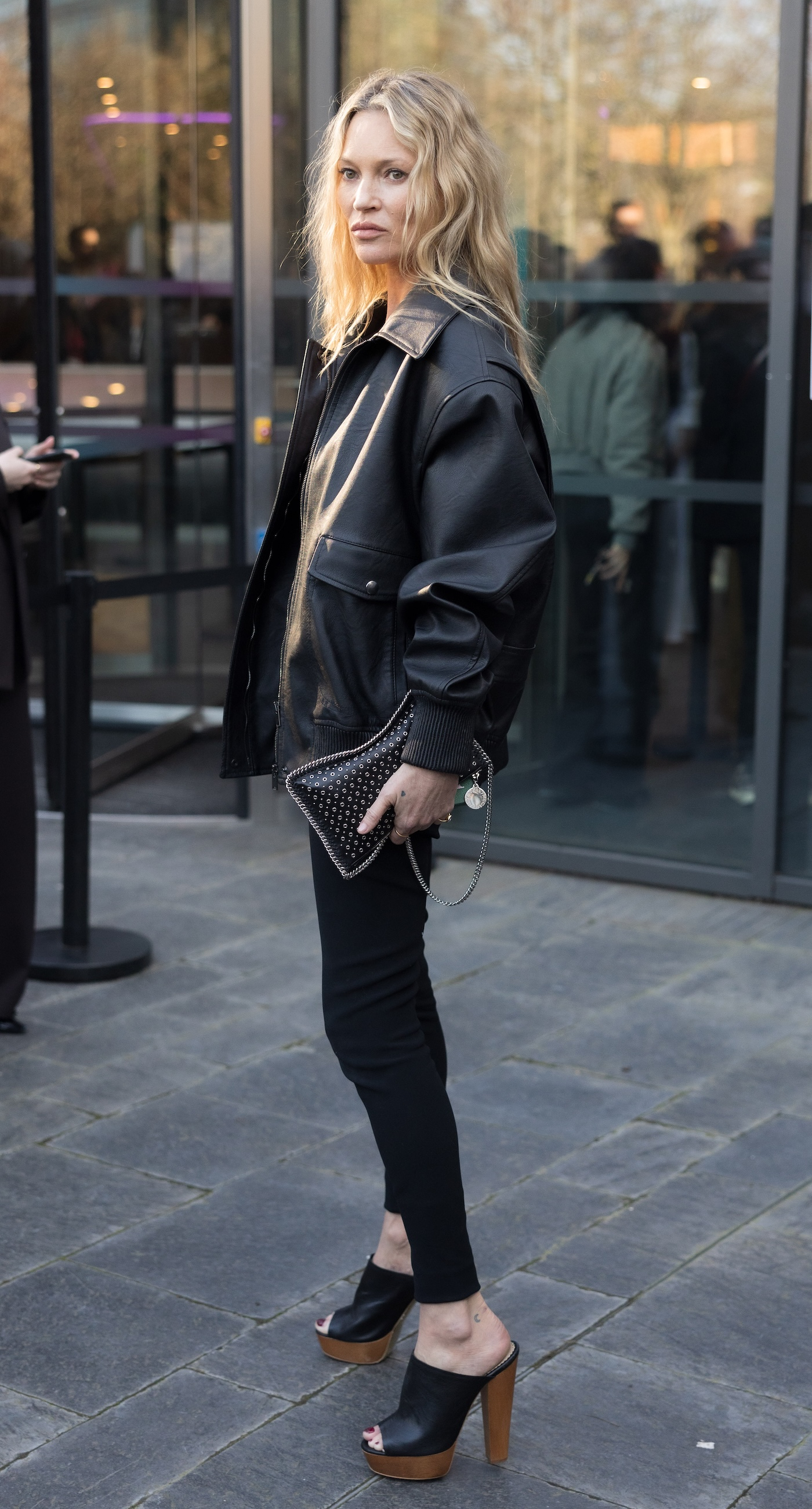 Kate Moss ma na sobie oversize'ową czarną bomberka z wegańskiej skóry marki Stella McCartney oraz obcisłe czarne skinny jeans, małą torebkę na łańcuszku oraz klapki mule na bardzo wysokim obcasie.