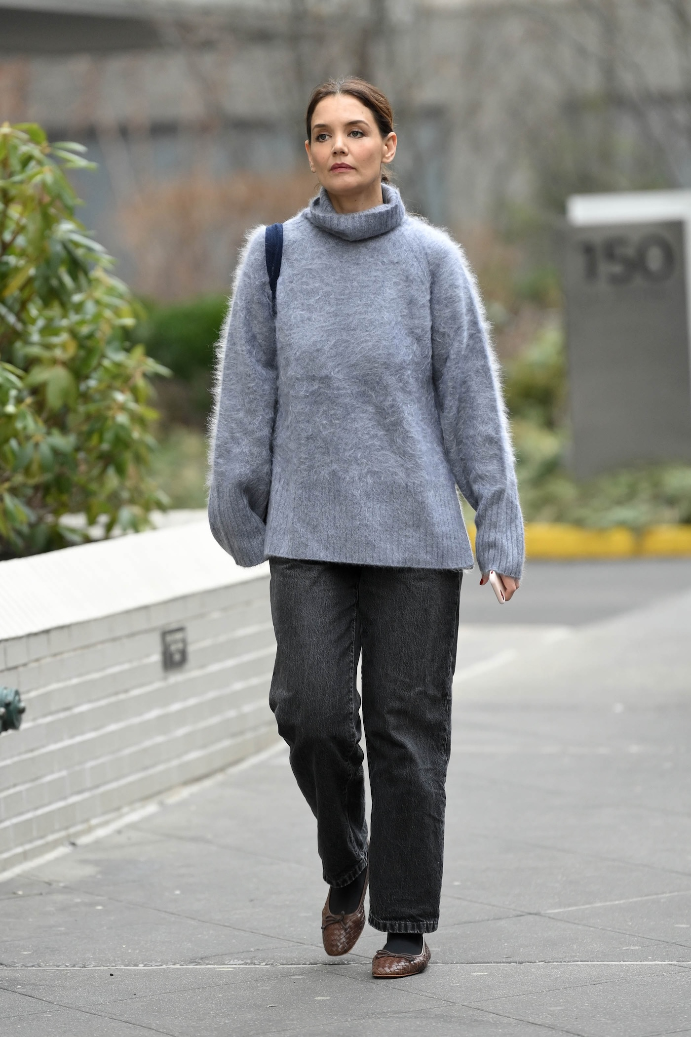 Katie Holmes podczas spaceru ulicami Londynu ma na sobie szary golf o kroju oversize, ciemnoszare baggy jeans, brązowe plecione baleriny ze skóry marki Dunes London, ciemne rajstopy i ciemną torebkę.