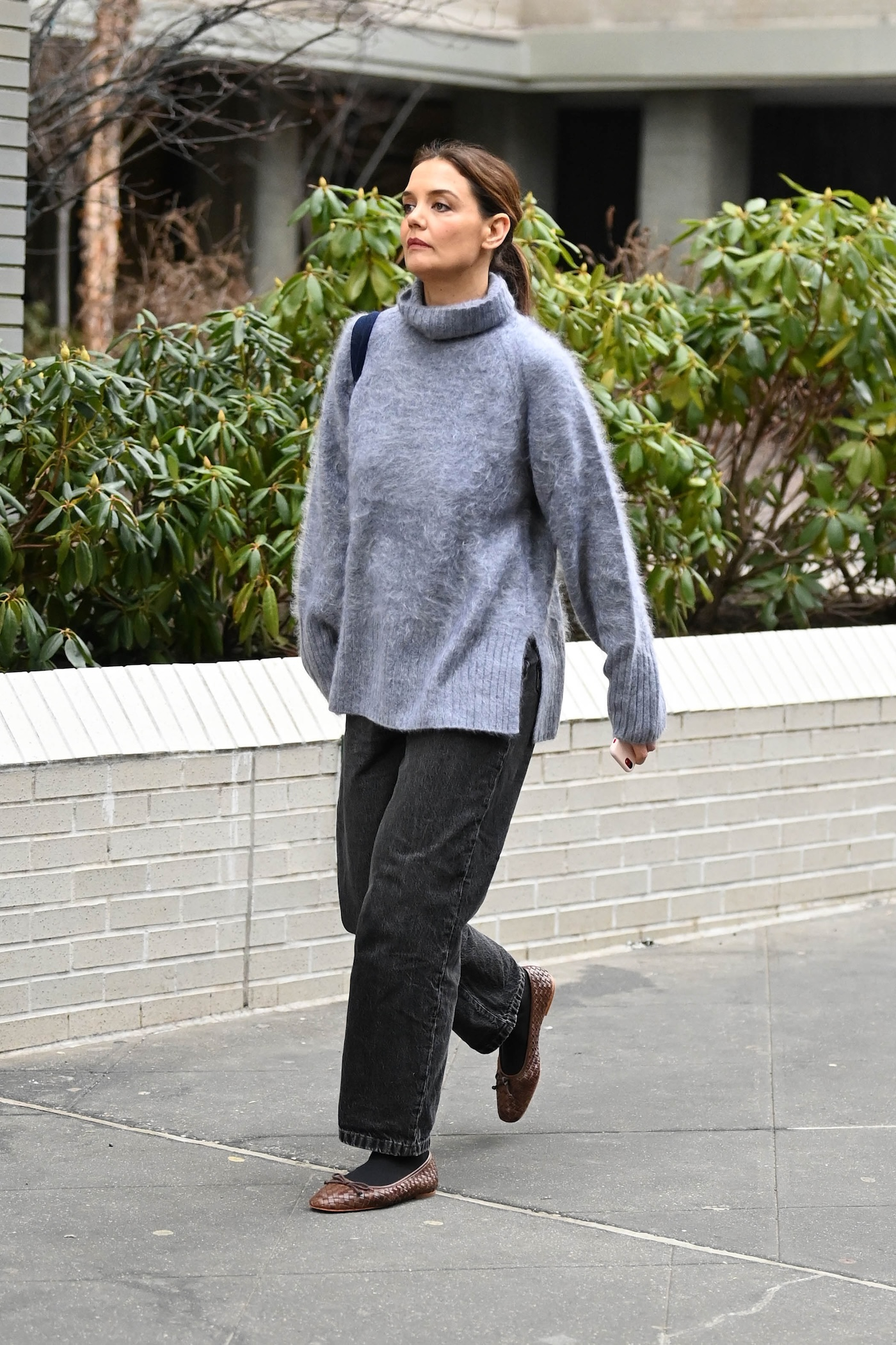 Katie Holmes podczas spaceru ulicami Londynu ma na sobie szary golf o kroju oversize, ciemnoszare baggy jeans, brązowe plecione baleriny ze skóry marki Dunes London, ciemne rajstopy i ciemną torebkę.