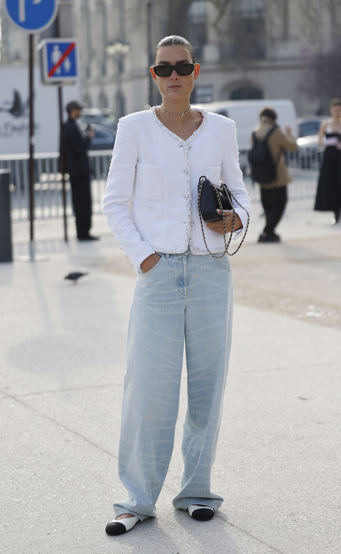 Kobieta sfotografowana podczas Paris Fashion Week, ma na sobie białe baleriny z czarnym noskiem, jasne jeansy z szeroką nogawką, biały damski żakiet bez klap Chanel, czarną torebkę Chanel na łańcuszku oraz czarne okulary przeciwsłoneczne.