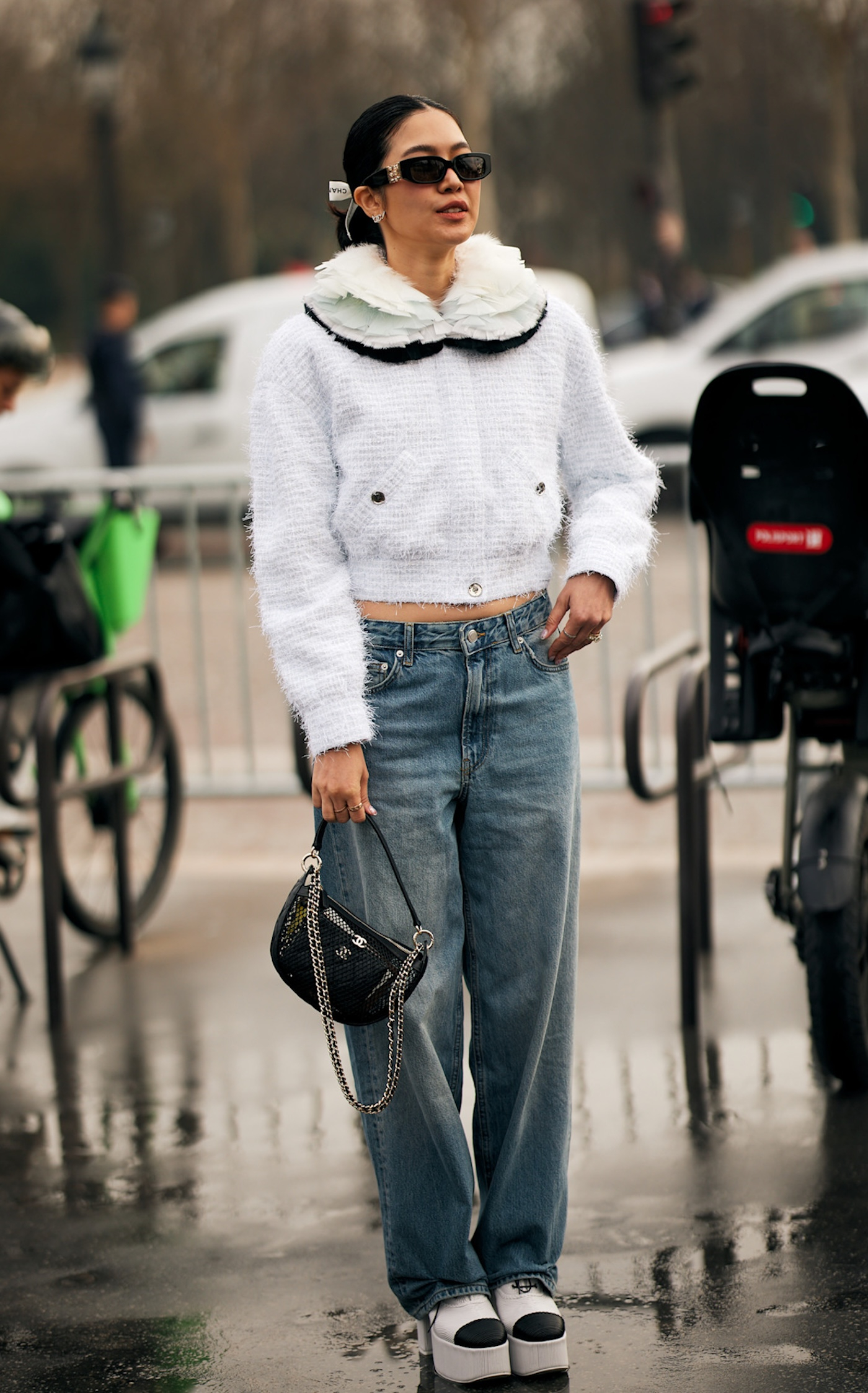 Kobieta sfotografowana podczas Paris Fashion Week, ma na sobie dwukolorowe buty na platformie, szerokie jeansy, tweedowy cardigan Chanel oraz czarną torebkę na łańcuszku.