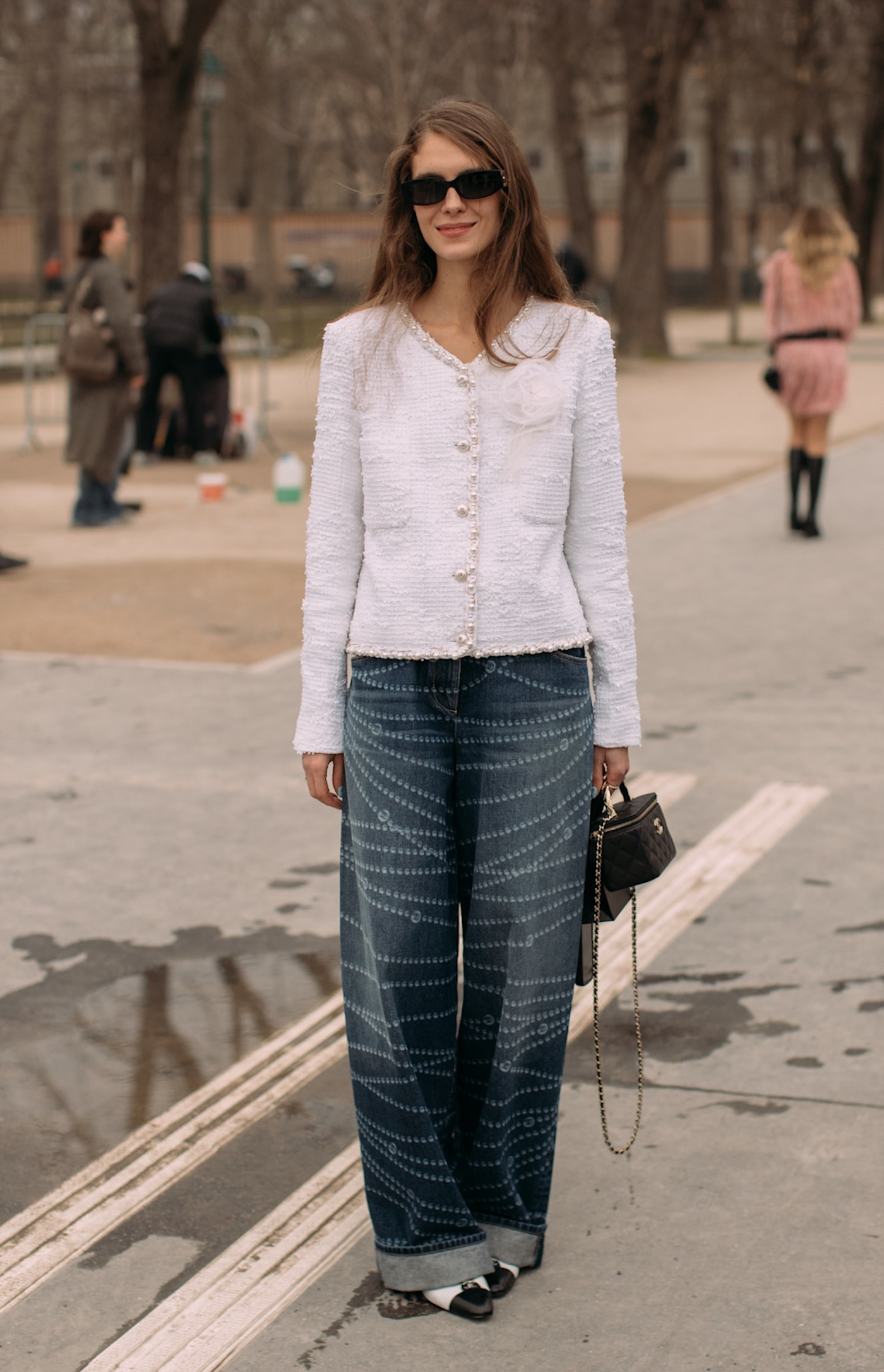 Kobieta sfotografowana podczas Paris Fashion Week, ma na sobie białe baleriny z czarnym noskiem, bogato zdobione baggy jeans, biały damski żakiet bez klap Chanel, czarną torebkę Chanel na łańcuszku oraz czarne okulary przeciwsłoneczne.
