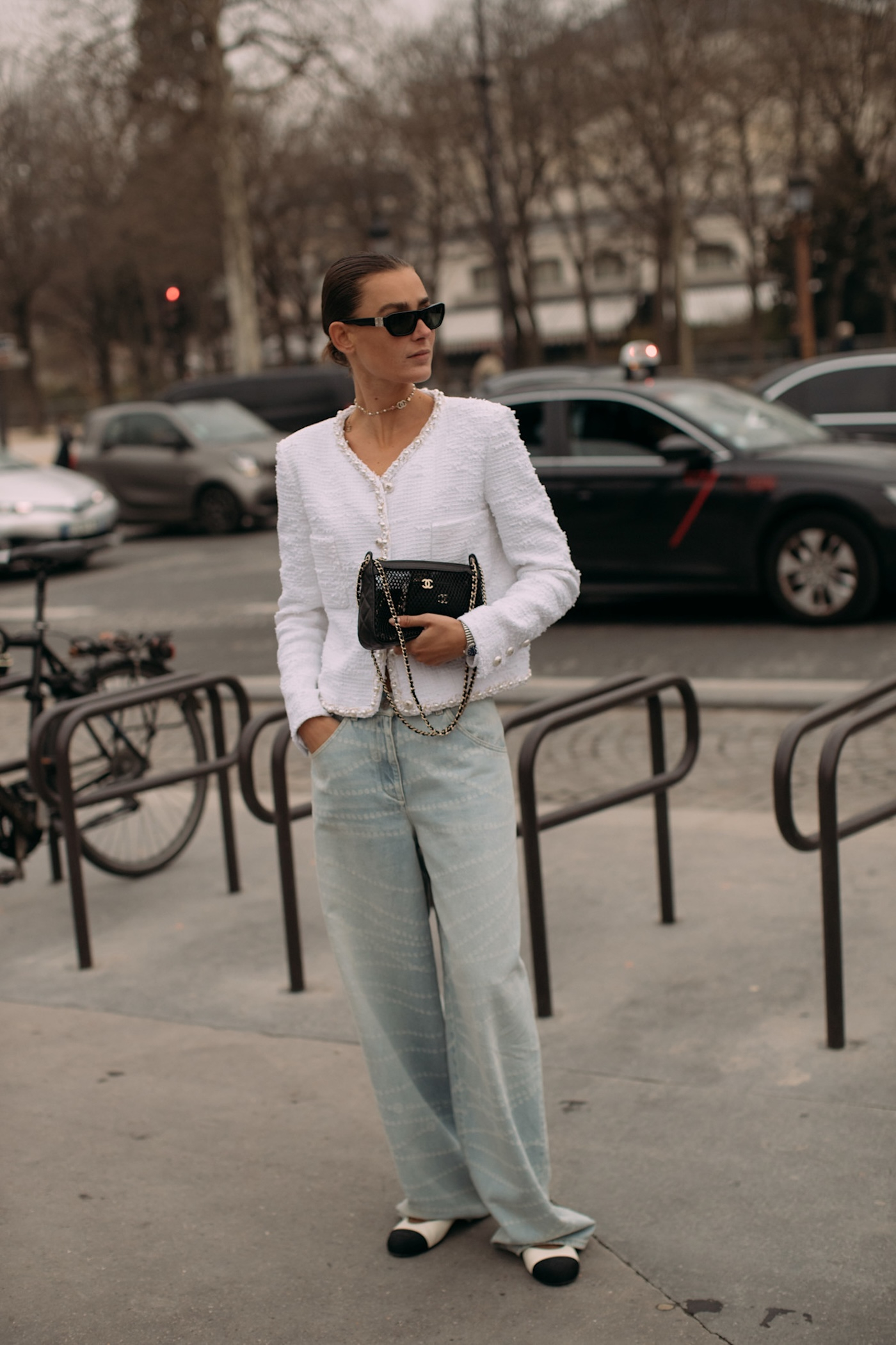 Kobieta sfotografowana podczas Paris Fashion Week, ma na sobie białe baleriny z czarnym noskiem, jasne jeansy z szeroką nogawką, biały damski żakiet bez klap Chanel, czarną torebkę Chanel na łańcuszku oraz czarne okulary przeciwsłoneczne.