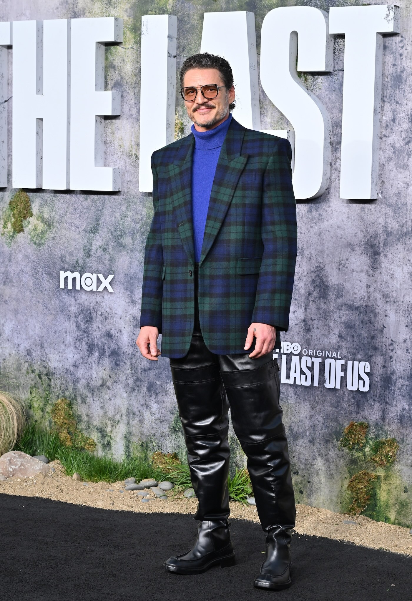 Pedro Pascal na premierze 2 sezonu serialu "The Last of Us" ma na sobie retro okulary z pomarańczowymi szkłami, golf w kolorze ultramaryny, marynarkę w kratę typu tartan, czarne skórzane spodnie oraz czarne bardzo wysokie buty oficerki z klamrami.