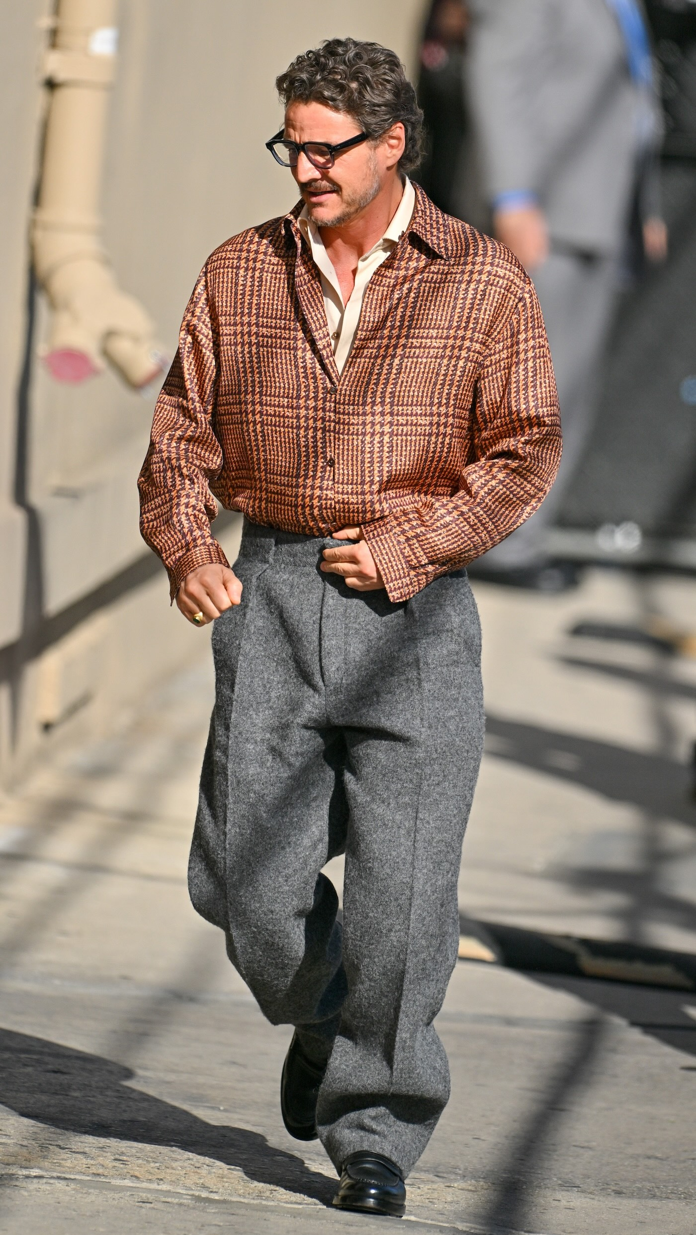 Pedro Pascal ma na sobie retro okulary, białą koszulę, kurtkę w brązową kratkę, szare garniturowe spodnie i czarne mokasyny.
