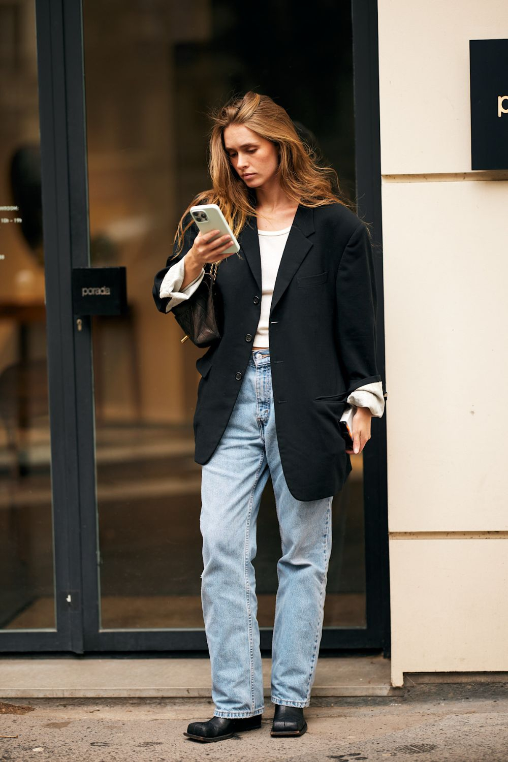 Kobieta w oversize'owej marynarce i boyfriend jeans, trzyma telefon, stojąc przed szklanymi drzwiami.
