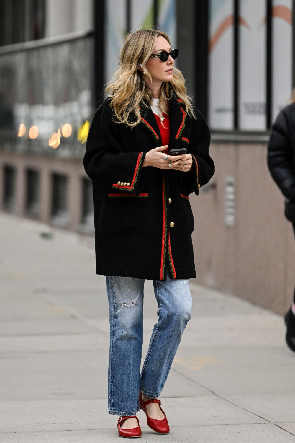 Kobieta w czarnym płaszczu i boyfriend jeans spaceruje po ulicy, trzymając telefon. Uzupełniają ją czerwone buty.